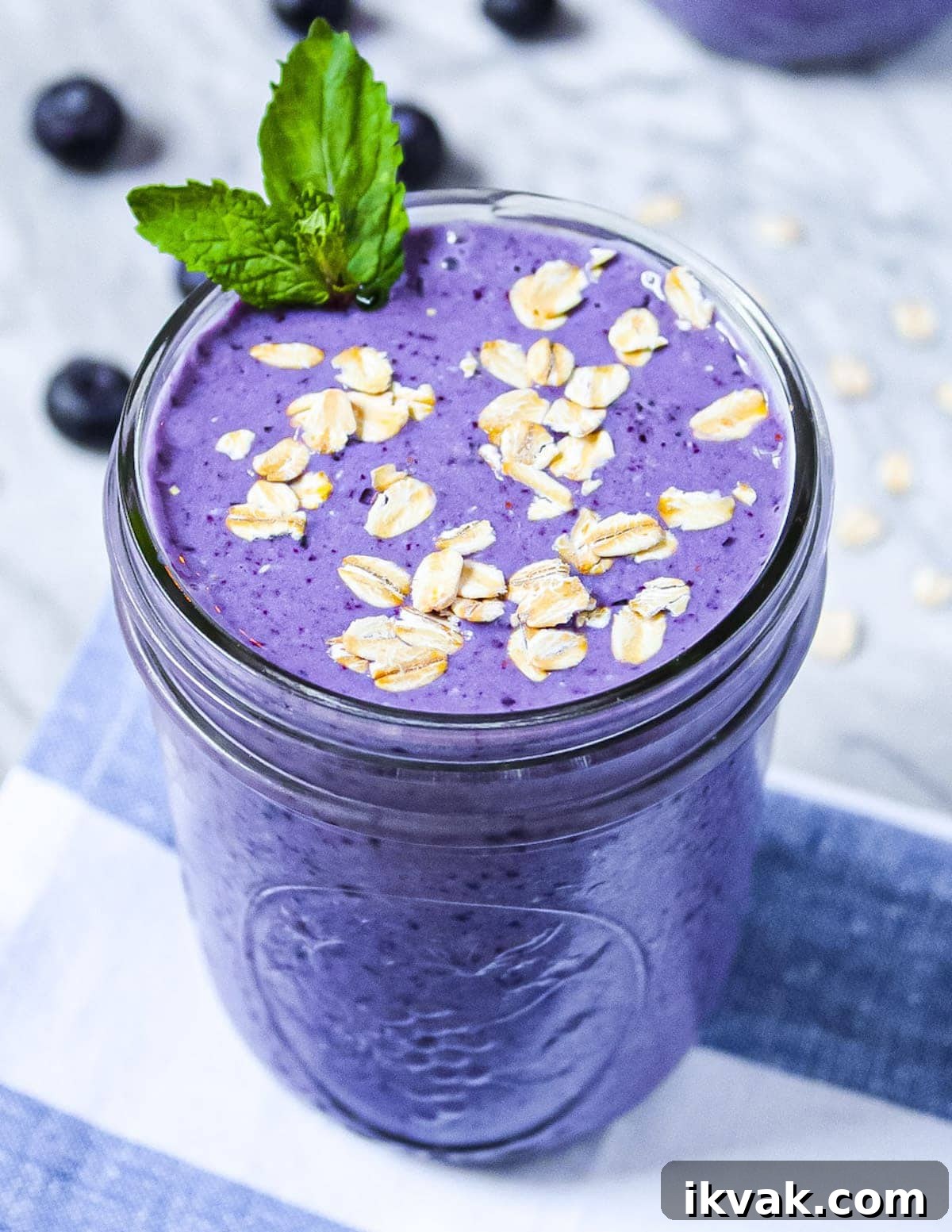 Blueberry Muffin Smoothie Recipe: Creamy Breakfast Shake 2 A purple smoothie garnished with green mint, uncooked oatmeal, sitting on a blue and white striped dish towel.