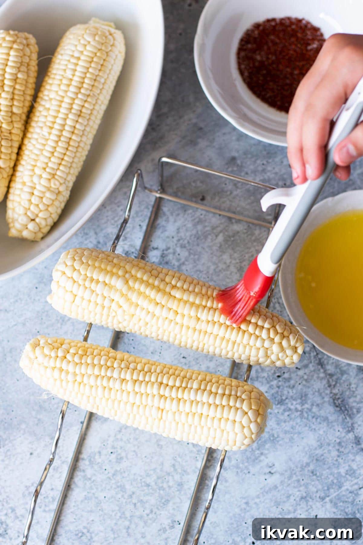 Person using basting brush to add butter to corn on the cob.