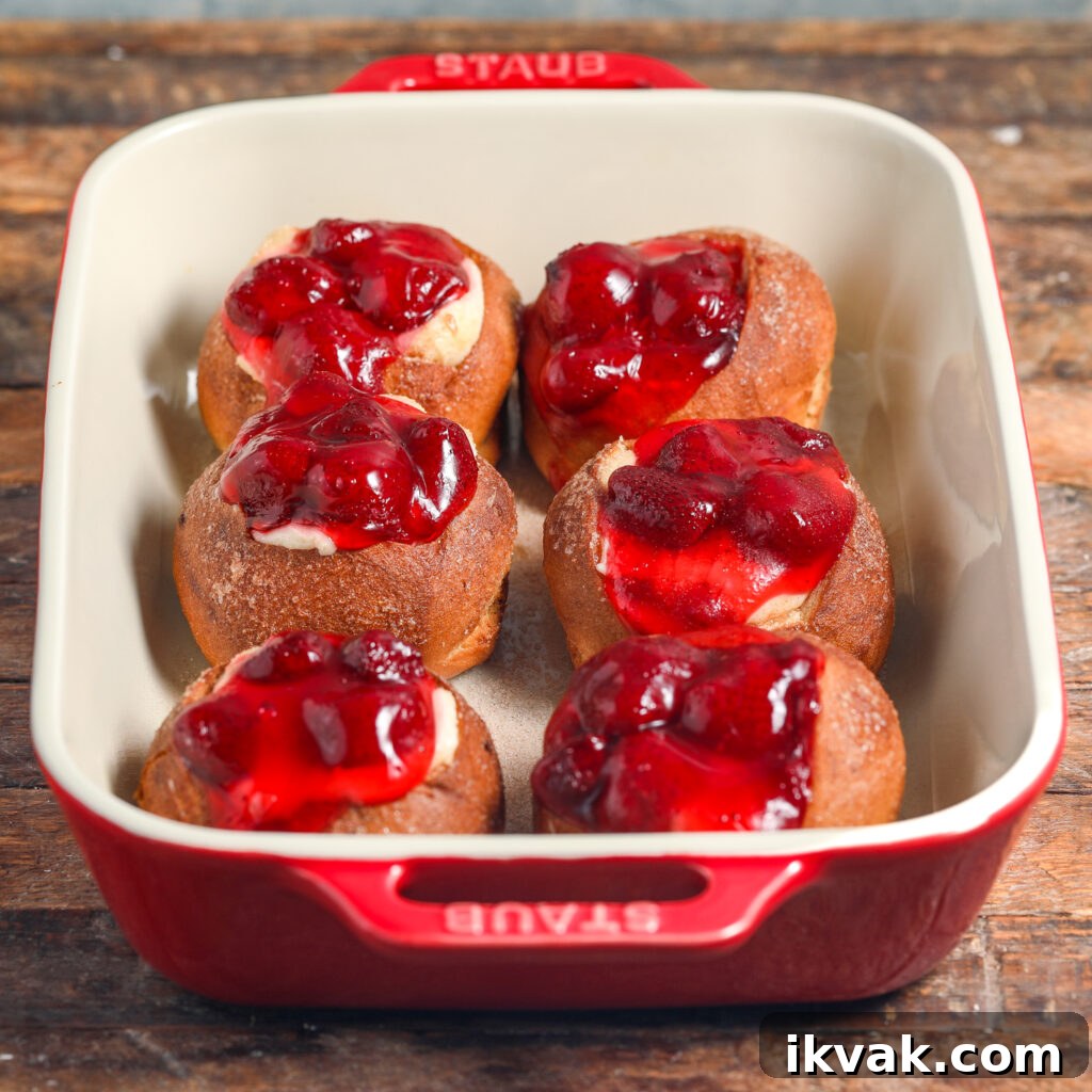 A red baking dish holds six round pastries, reminiscent of strawberry danish sliders, filled with cream cheese and topped with bright red cherry filling, sitting on a rustic wooden surface.