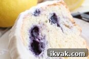 A slice of lemon blueberry bundt cake frosted with lemon cream cheese frosting sitting on a small white plate. Above the plate is a whole lemon and a blueberry lemon bundt cake frosted with lemon cream cheese frosting with a slice taken out of it.