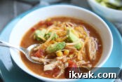 A white bowl full of chicken tortilla soup with a spoon in it sitting on a blue plate. The soup is topped with grated cheese and pieces of avocado.