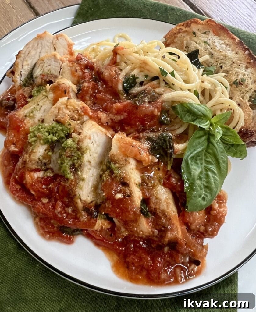 Sliced Pomodoro Chicken on white dinner plate with spaghetti, bread, basil garnish and a little pesto.