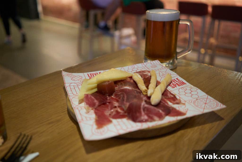A plate of jamón iberico and manchego cheese with breadsticks on a table with a beer in the background.