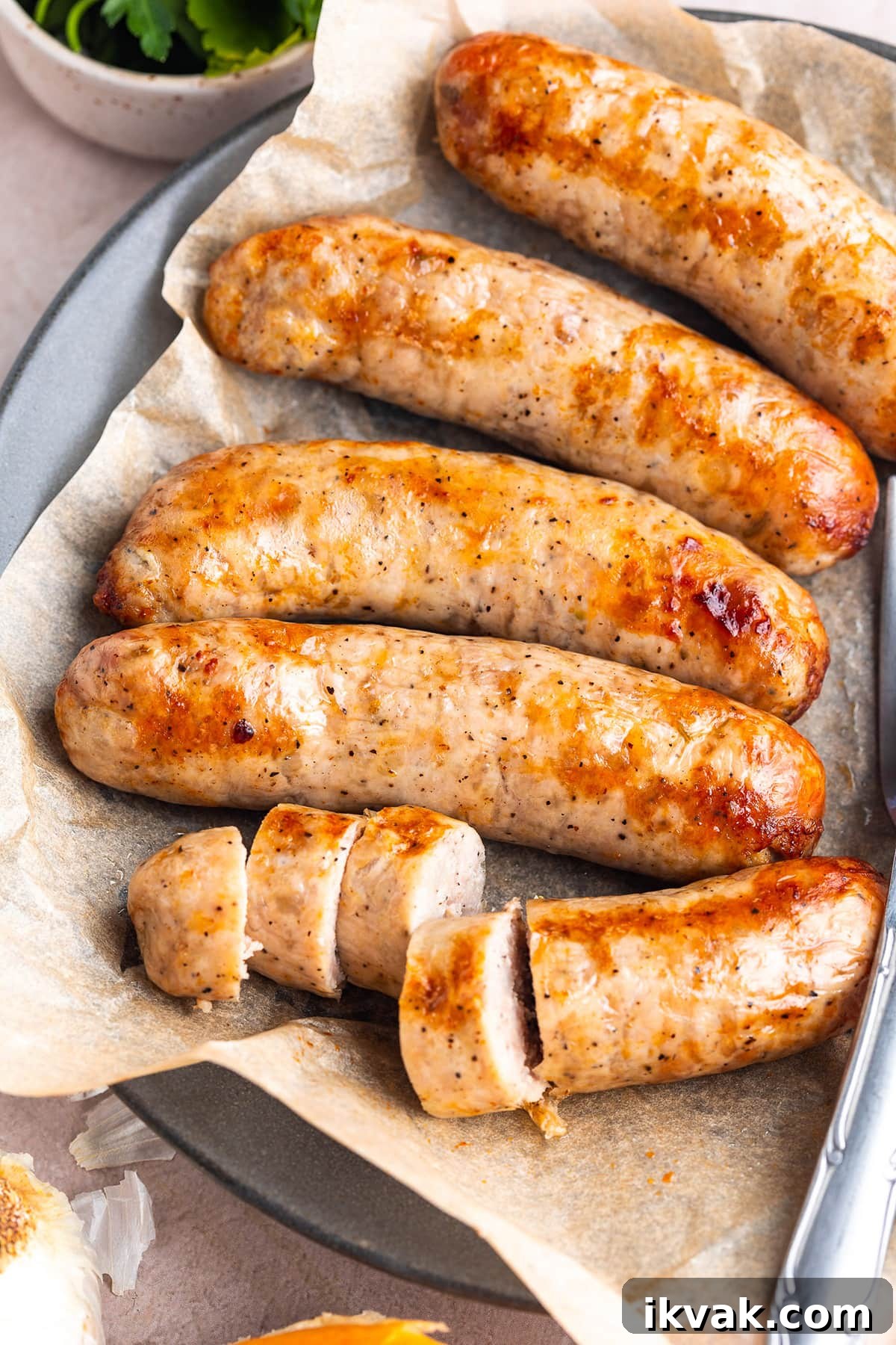 5 Italian sausages on a plate lined with parchment paper. One of the sausages is sliced into thick discs.