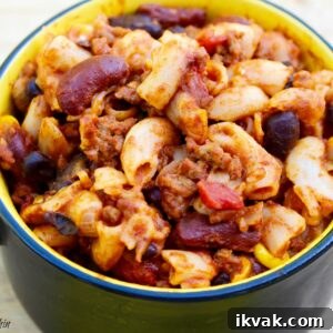 A hearty bowl of chili mac combines macaroni pasta, kidney beans, ground meat, and diced tomatoes in a rich, spicy sauce. The dish is served in a yellow and black handled bowl, placed on a light wooden surface.
