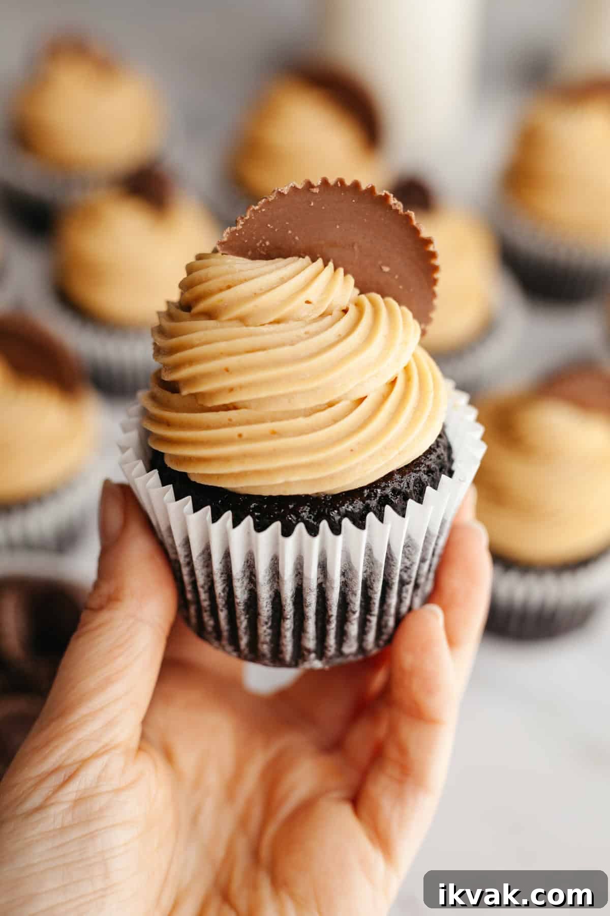 A hand holding a chocolate peanut butter cupcake with a Reese's cup on it.