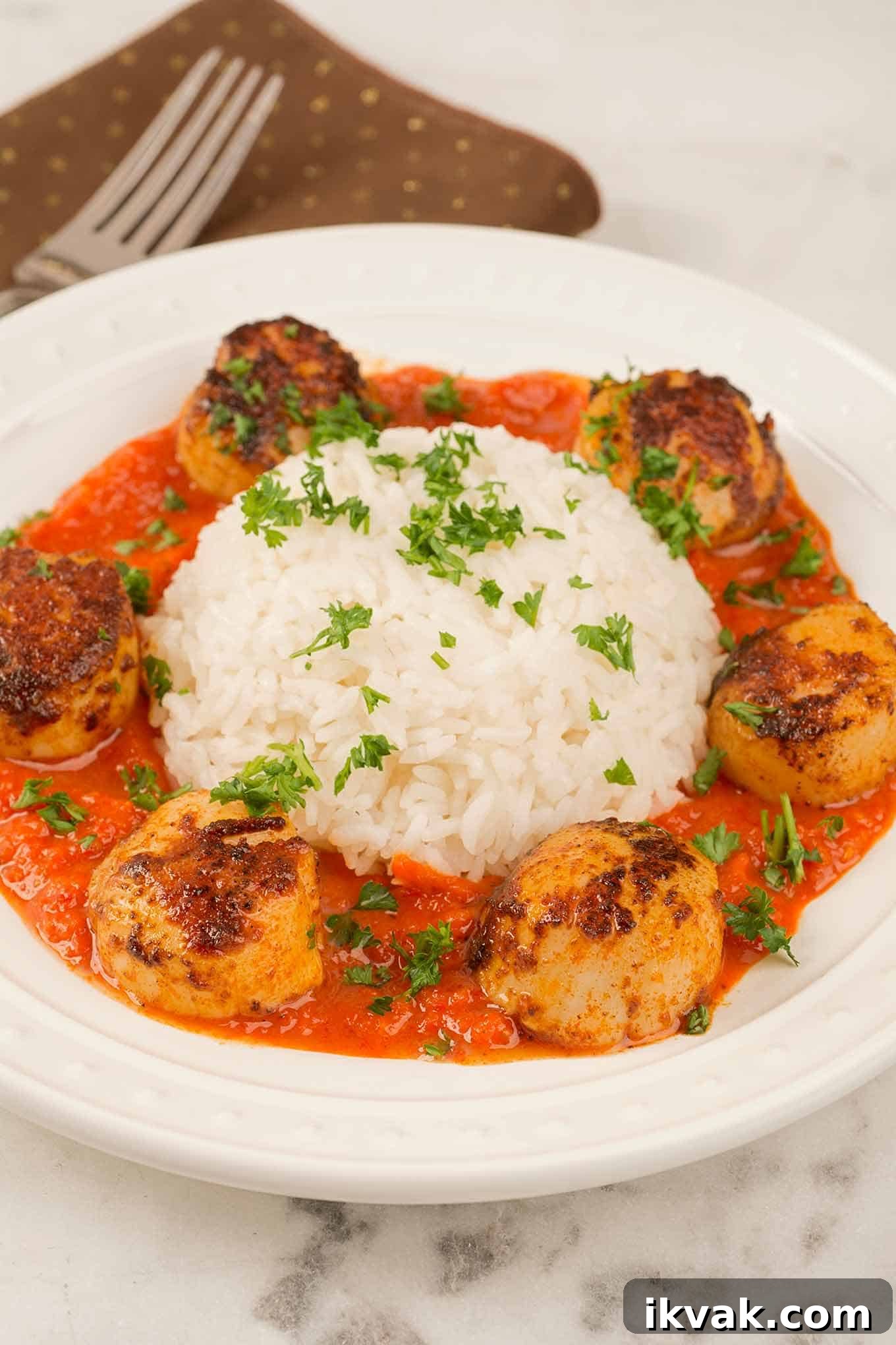 A plate of paprika-crusted, pan-seared sea scallops in roasted red pepper sauce arranged around a serving of white rice, all garnished with chopped parsley.