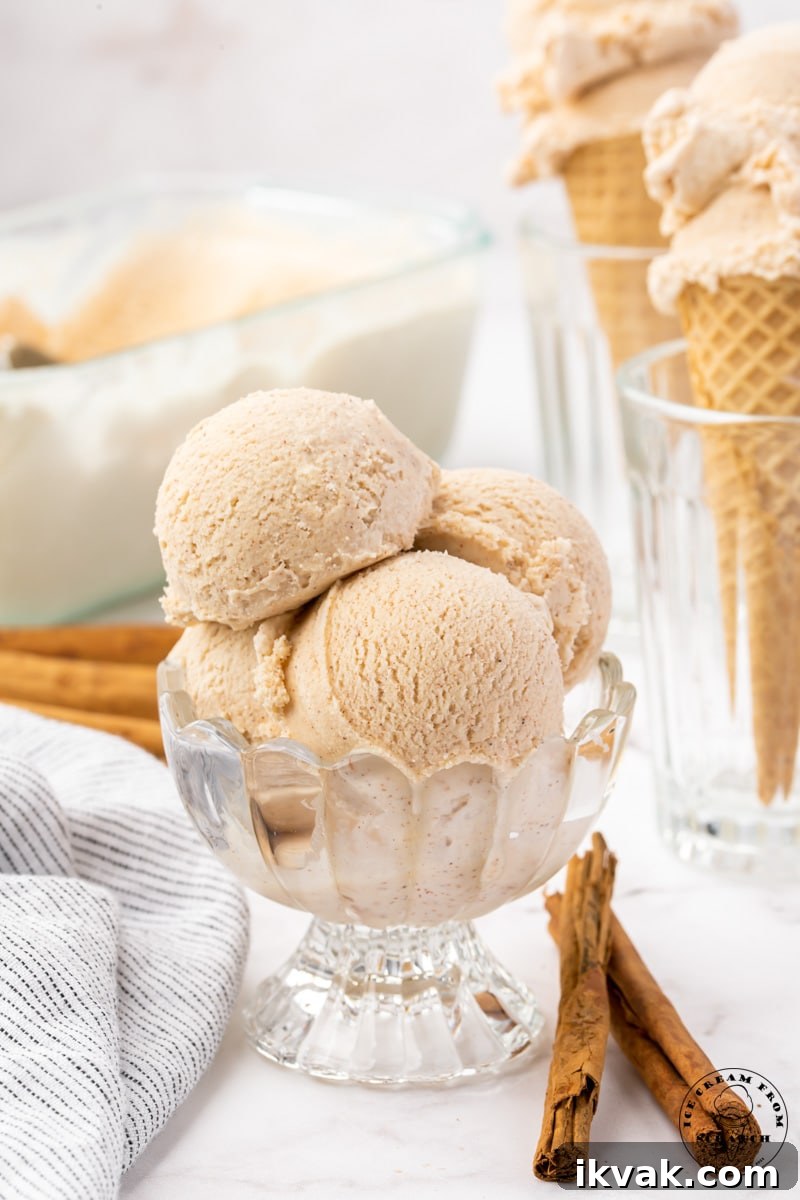 four scoops of cinnamon ice cream in a footed glass dish. Cinnamon sticks are on the counter in front of the dish. Two cones filled with ice cream are in the background.