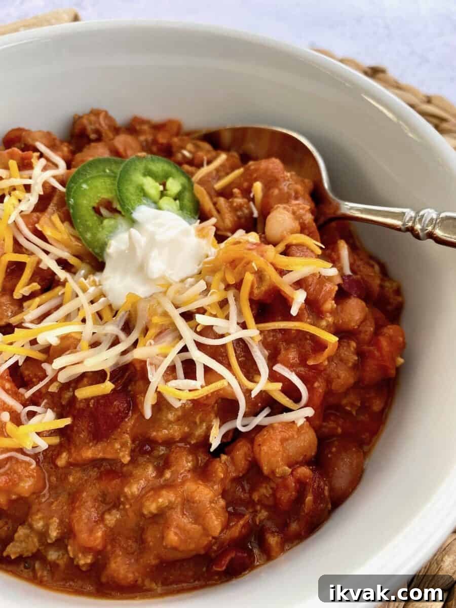A bowl of chili with shredded cheese, sour cream and jalapenos on top with a spoon on the side