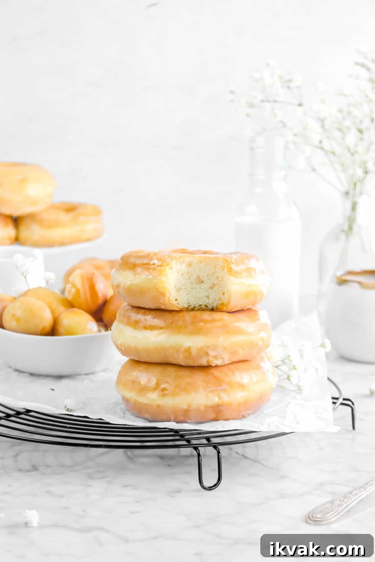three doughnuts stacked with a bite taken out of one, flowers, milk, and more doughnuts behind