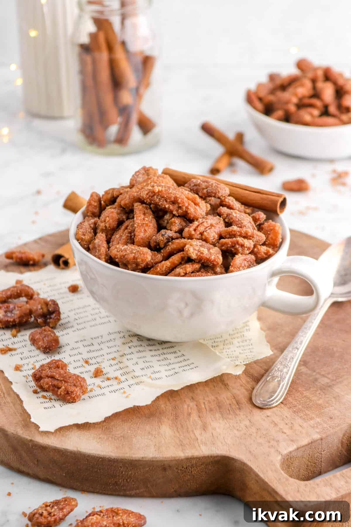candied pecans in white mug with fairy lights, cinnamon sticks, and more pecans scattered around