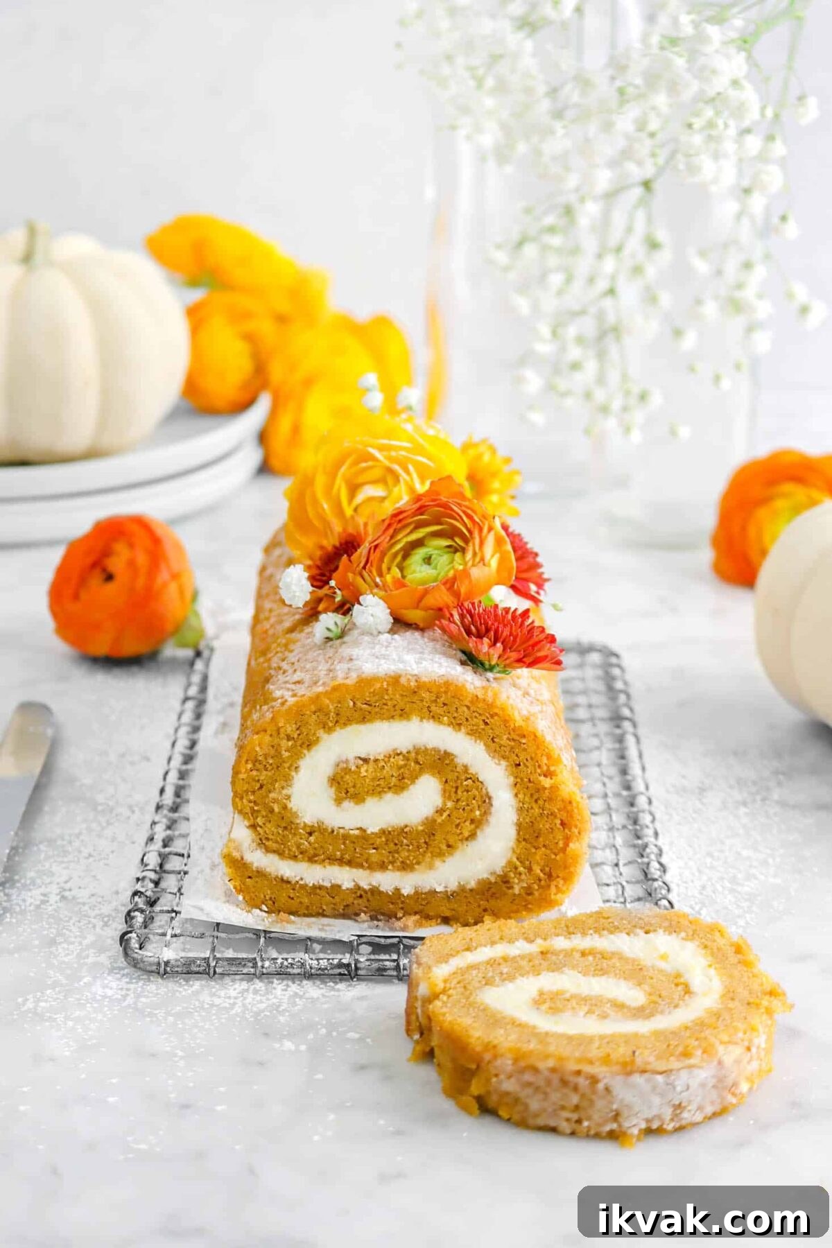 pumpkin roll on wire rack with small piece of parchment, flowers, on top, more flowers around, and two white pumpkins