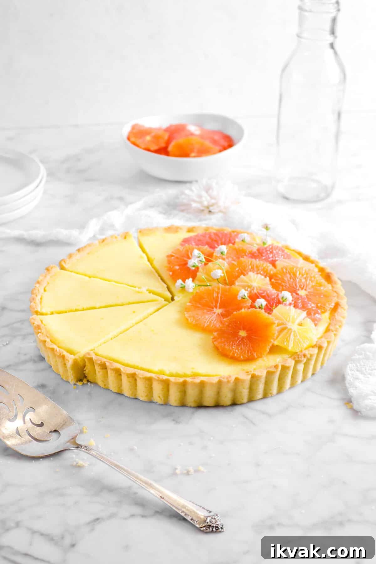 citrus tart with three slices laying next to it, a cake knife, white napkin, glass, and bowl of citrus slices