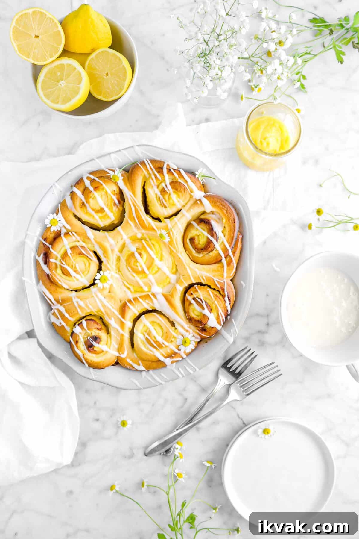 overhead shot of lemon rolls in pie pan with a napkin, forks, lemons, flowers, lemon curd, and plates