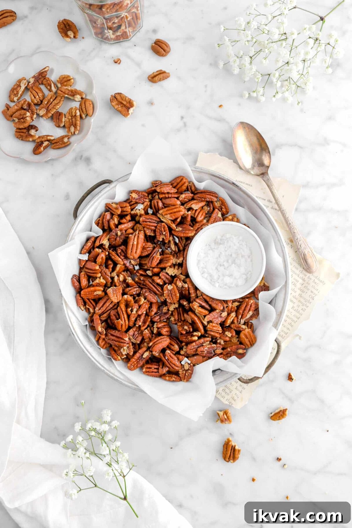 pulled back shot of roasted pecans in old cake pan with parchment paper, a bowl of salt, vintage spoon , flowers, book pages, and other pecans around