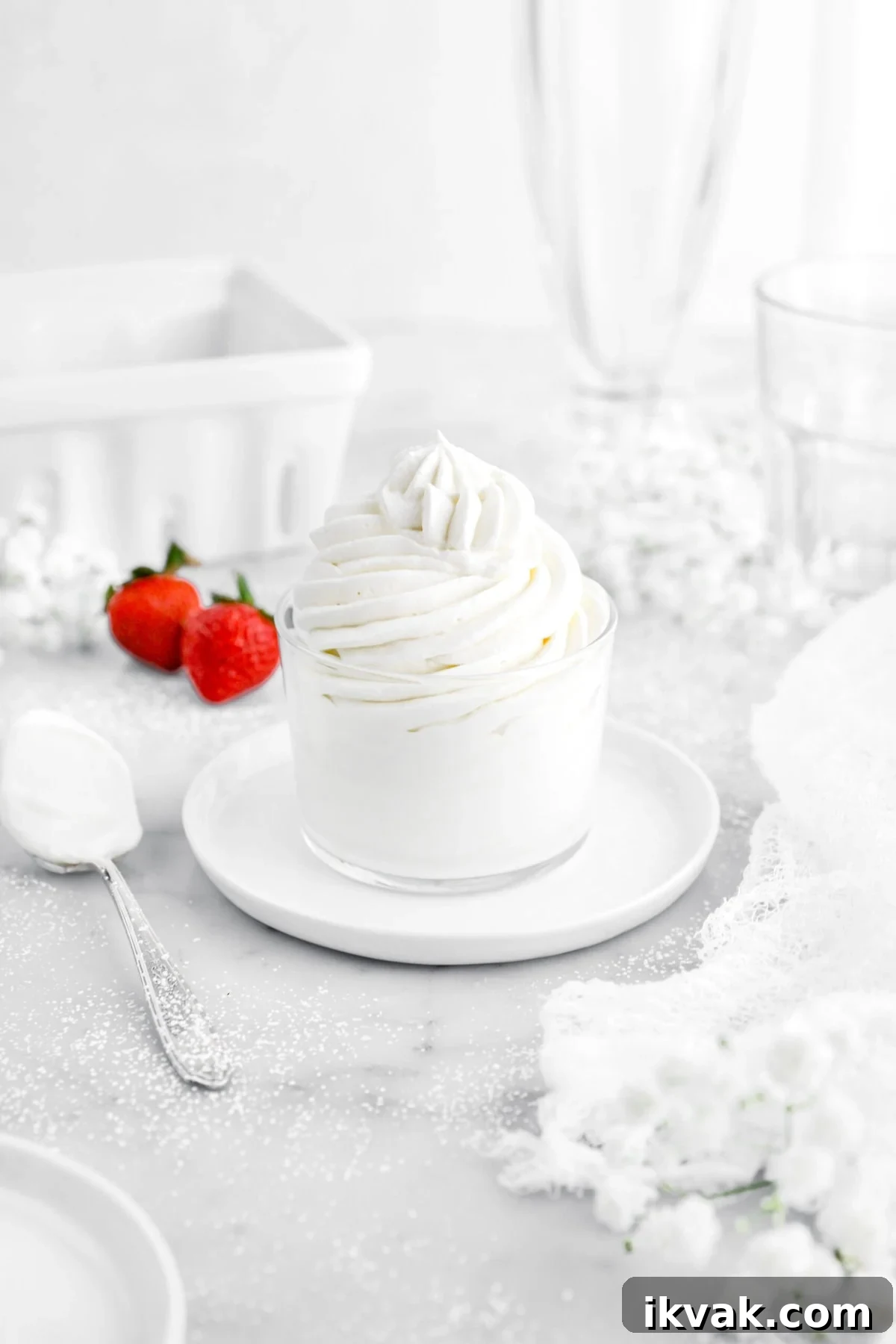 angled image of piped chantilly cream in small glass on top of white plate with spoon beside and two strawberries behind.