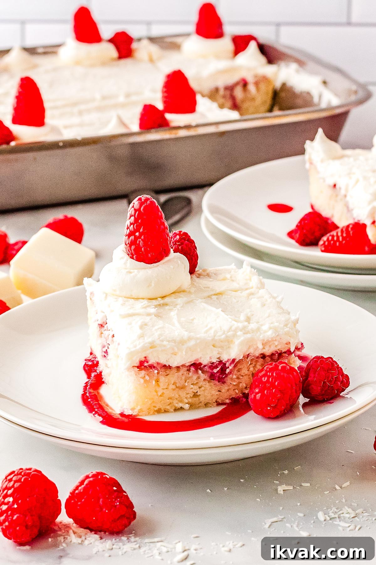 A slice of white chocolate and raspberry cake on a plate garnished with raspberries, puree, and white chocolate shavings.