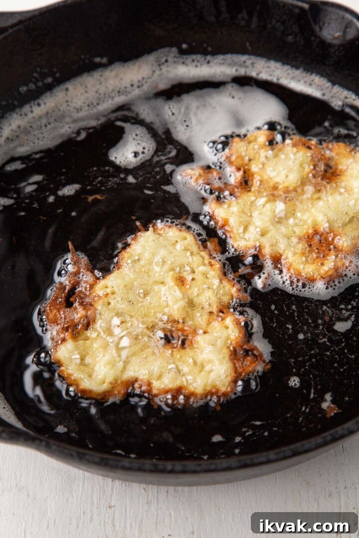 Crispy Potato Pancakes Recipe: Golden Hash Browns from Scratch 9 potato pancakes frying in oil in a cast iron skillet, raw on top.
