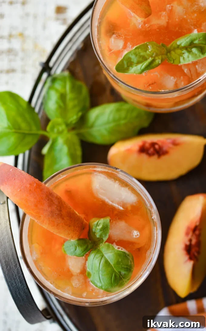 Two peach bourbon cocktails on a round wooden tray with fresh peaches and fresh basil.