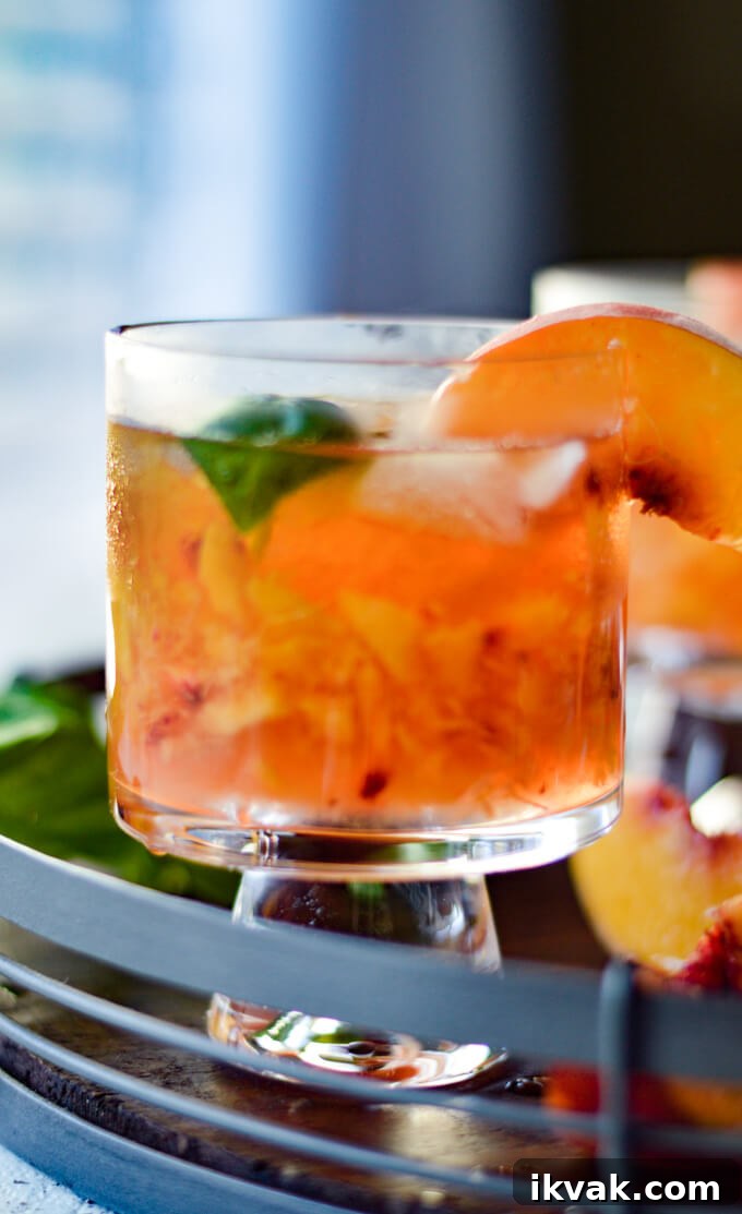 A front view of a peach bourbon cocktail on a round wooden tray with fresh peach slices and fresh basil scattered around.