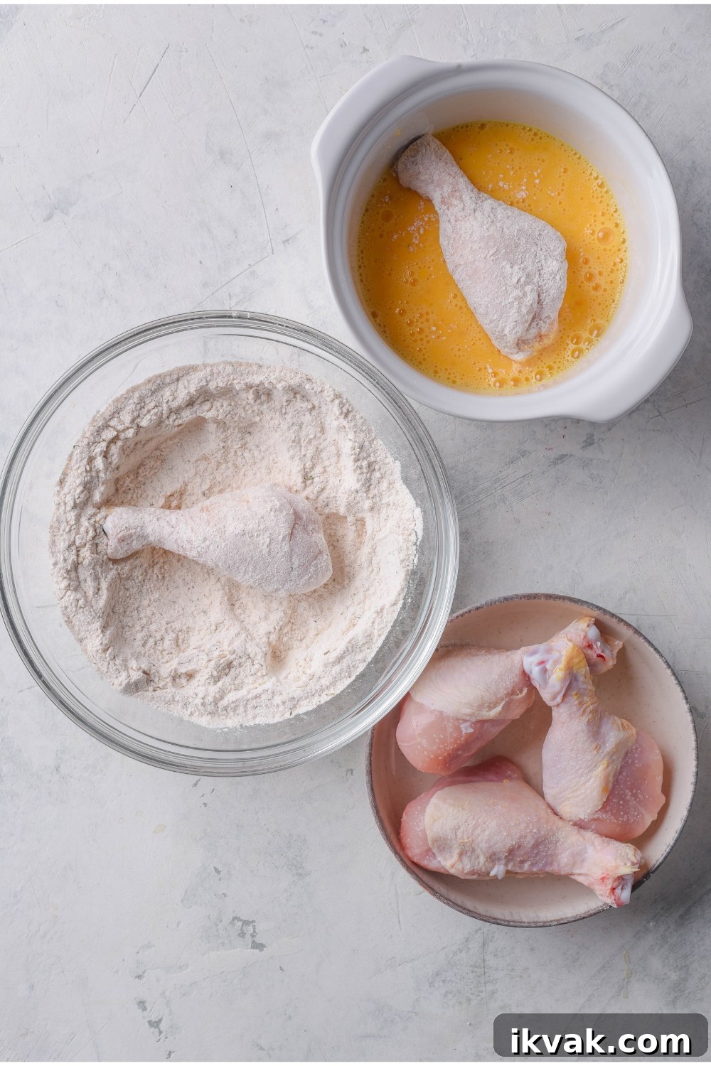 Three bowls of raw chicken, a dry flour mixture with a piece of chicken in it, and a bowl of beaten eggs with a piece of flour-coated chicken in it.