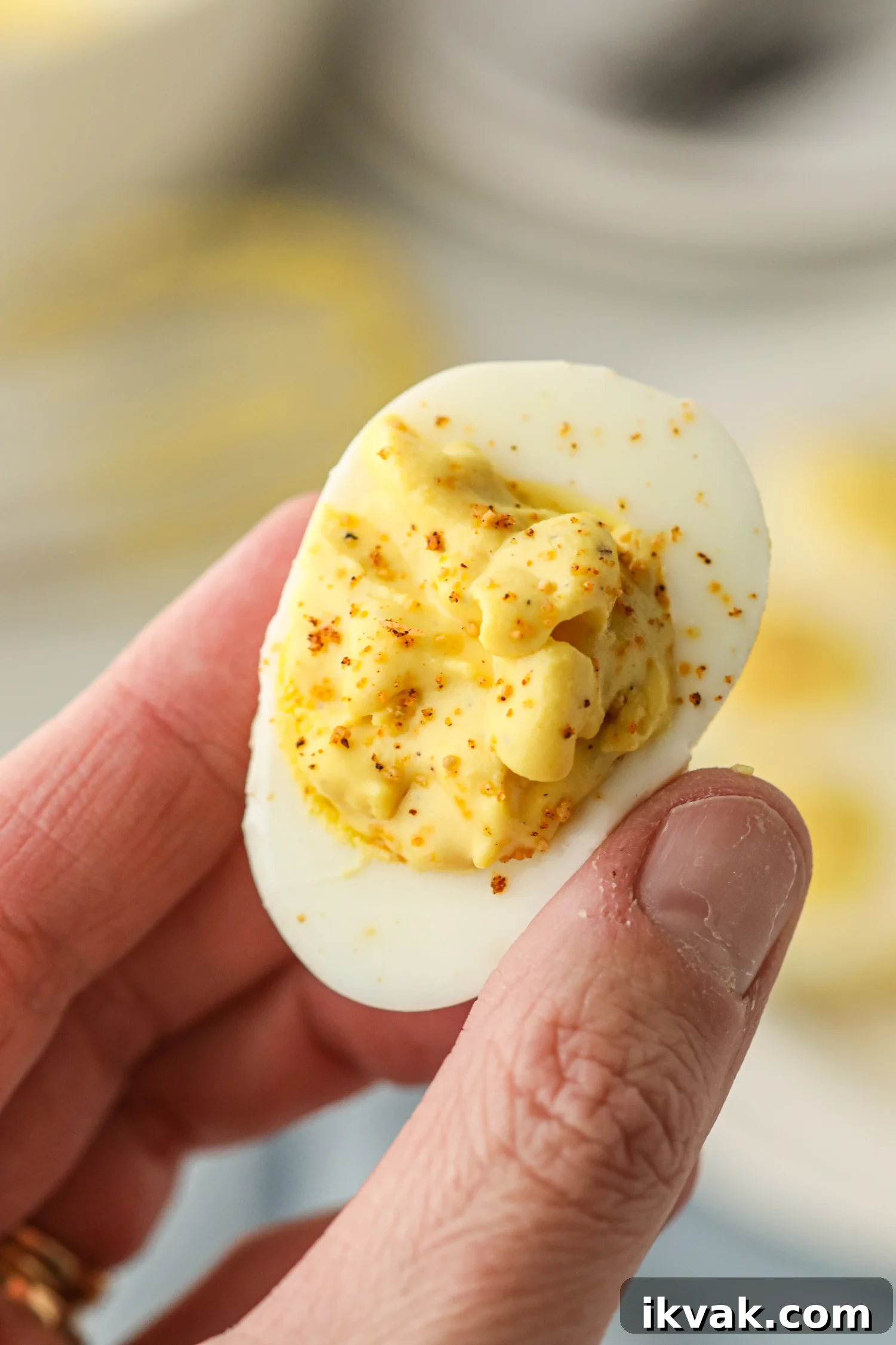 A close-up of a hand holding a smoked deviled egg. The egg white is filled with a creamy, yellow yolk mixture and sprinkled with seasoning. The texture of the filling appears smooth with a slight rustic consistency. The background is blurred, showing more deviled eggs on a plate.