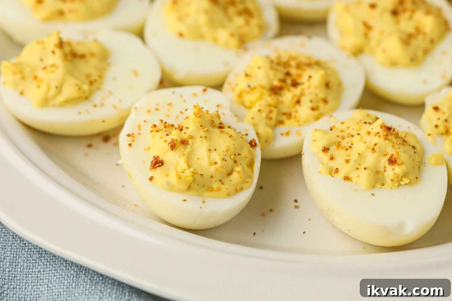 A close-up of smoked deviled eggs arranged on a white plate. Each egg half is filled with a creamy, yellow yolk mixture and topped with a light sprinkling of seasoning. The eggs have a slightly smoky tint, and the filling appears fluffy and well-mixed. The background includes a soft blue napkin, adding a touch of contrast to the presentation.