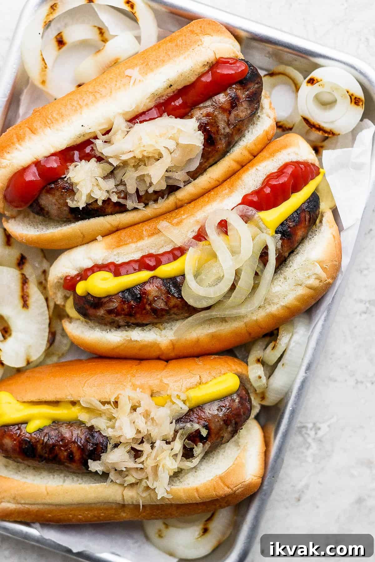 Grilled brats in buns on a metal tray with some grilled onions.