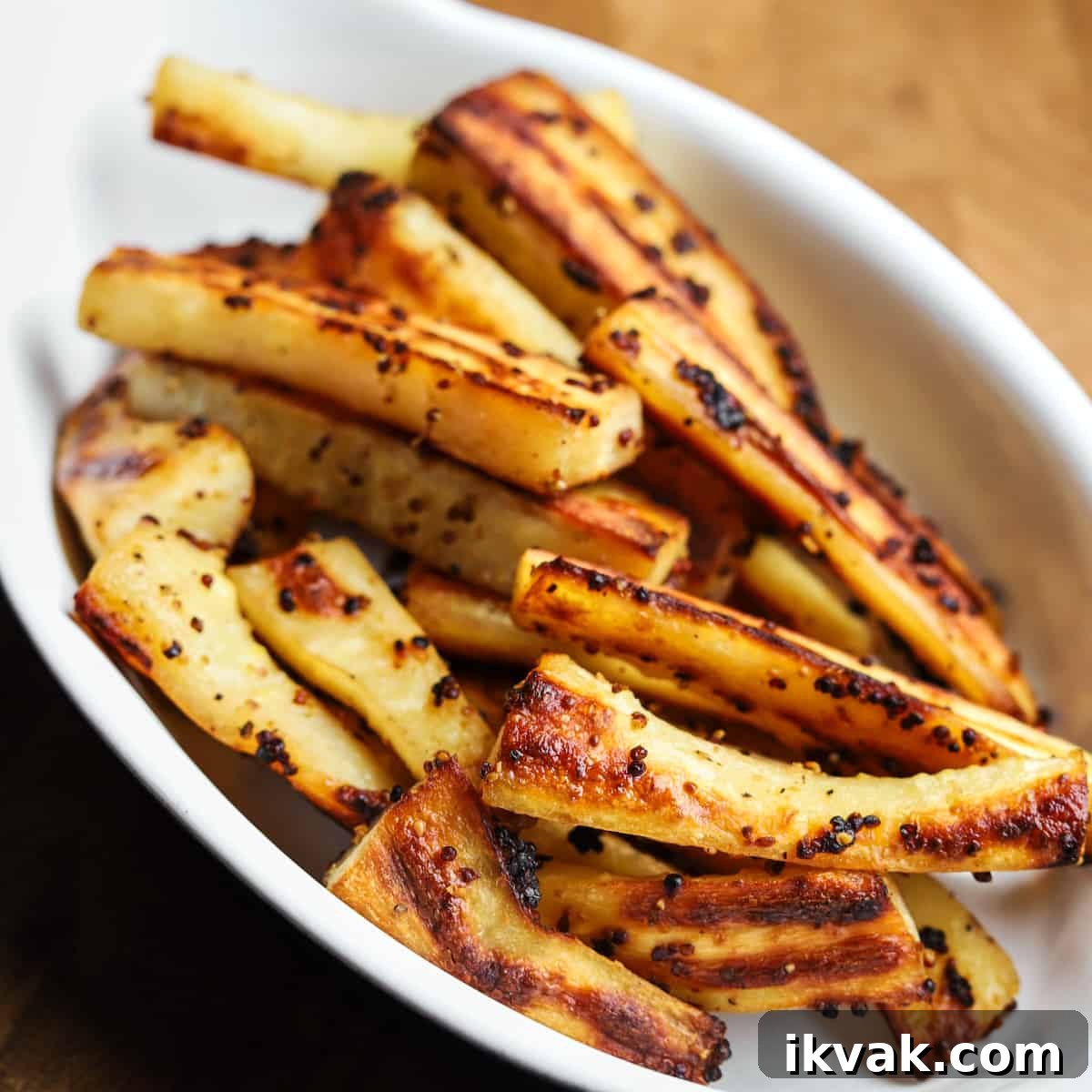 Square photo of roast parsnips in a white oval oven dish placed on a wooden table.
