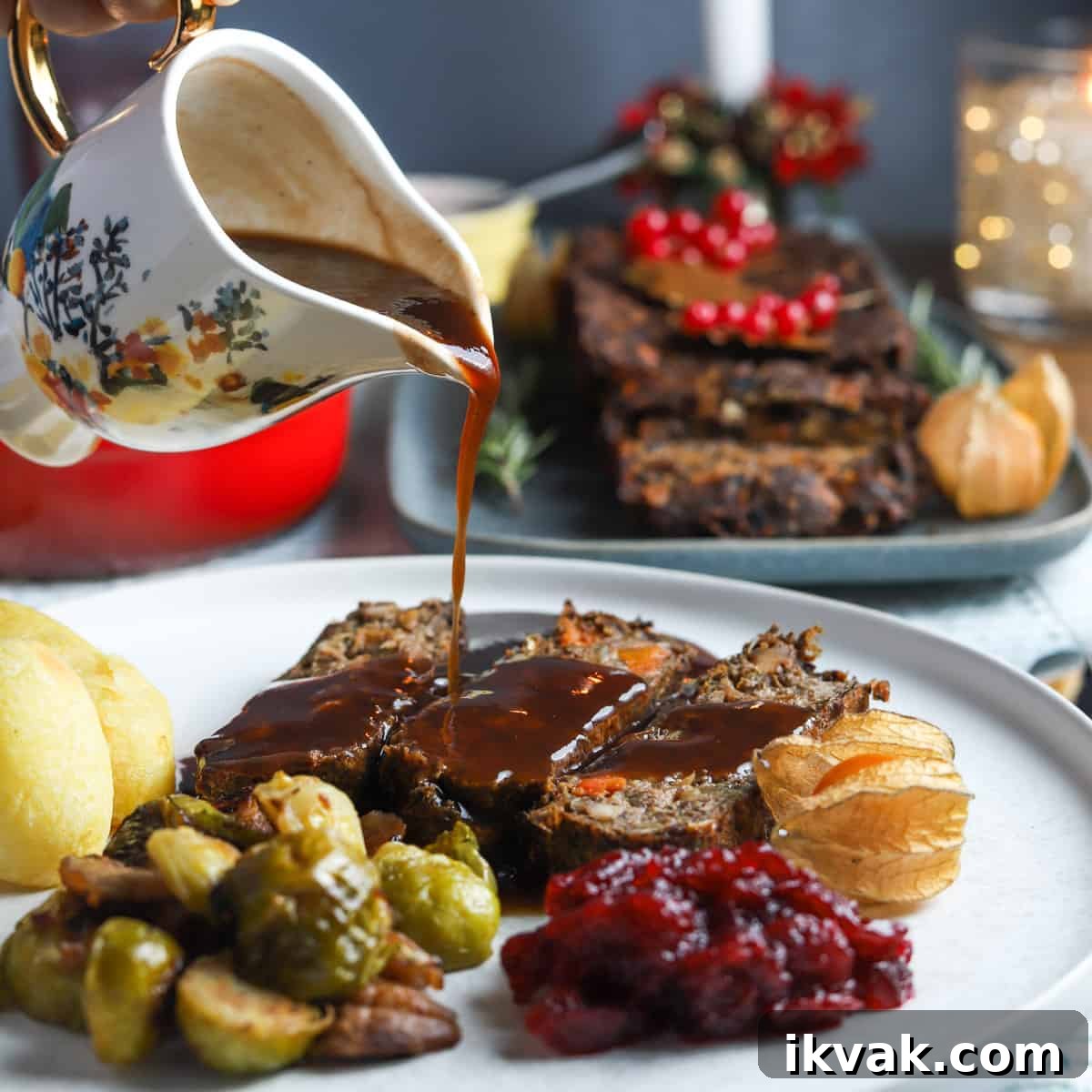 Photo of a plate filled with 3 slices of this vegan nut roast, some roast potatoes, sprouts, cranberry sauce, and a pretty jug pouring gravy over the top, with the full vegan nut roast in the background.