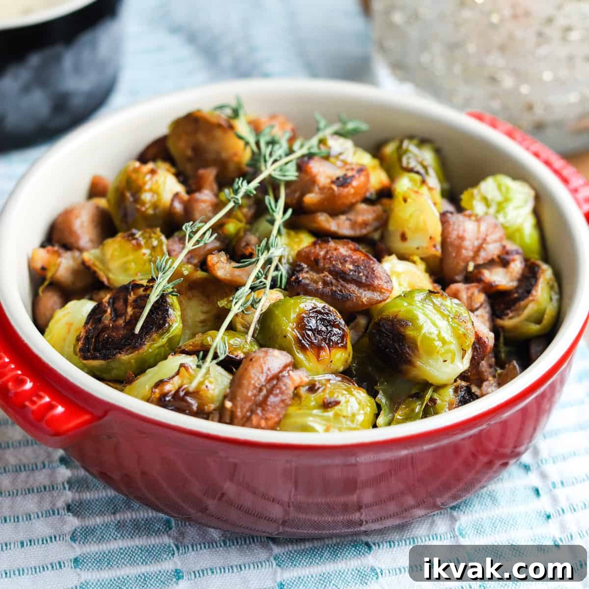 Photo of Christmas roast brussels sprouts with chestnuts in a red bowl on a tablecloth with tablescape in the background.