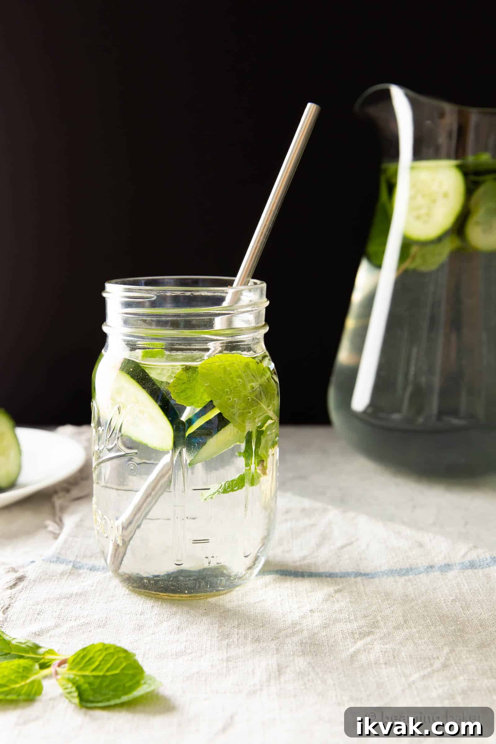 Glass of cucumber mint water with a metal straw inside, and pitcher in the background