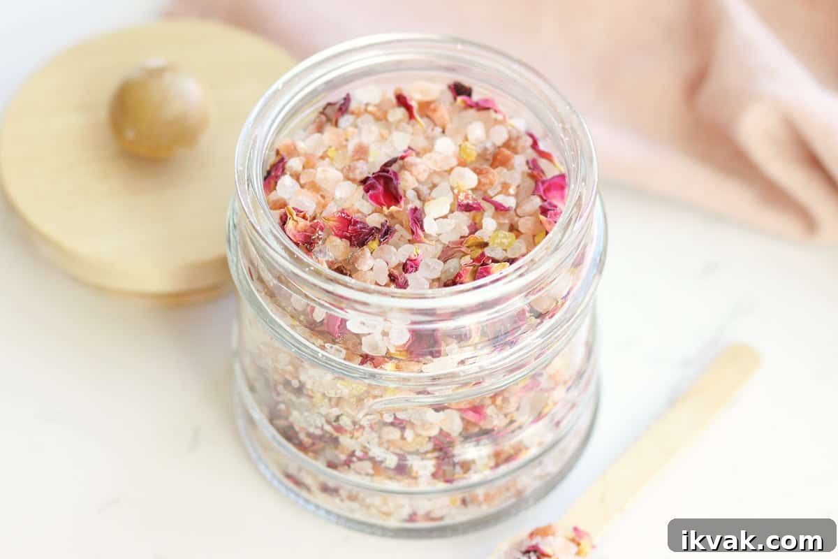 a small glass jar with a wooden lid and small wooden spoon full of rose bath salts with rose petals in it.