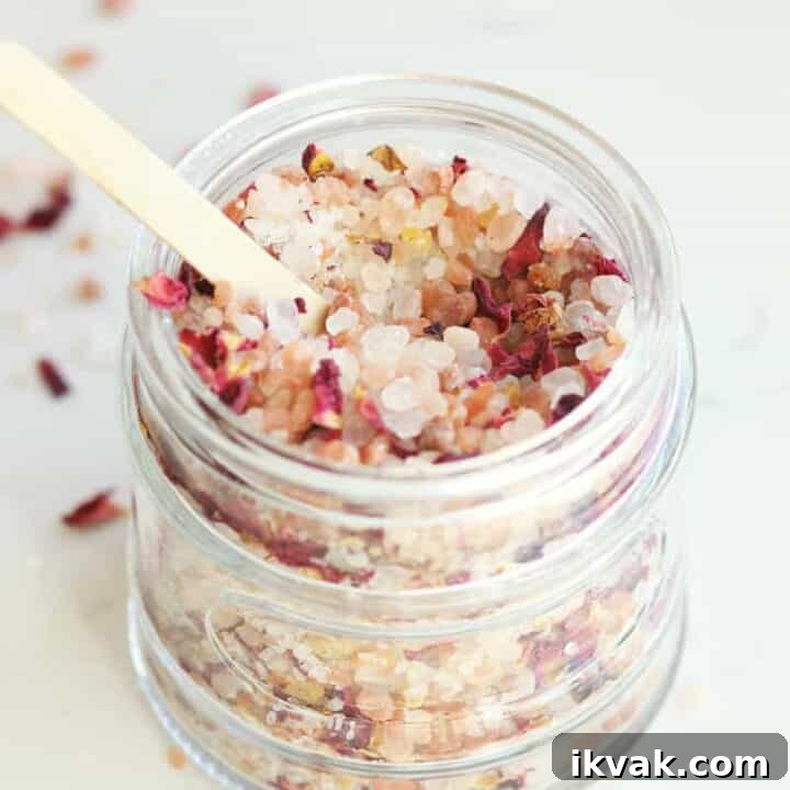 a small glass jar with a wooden lid and small wooden spoon full of rose bath salts with rose petals in it.