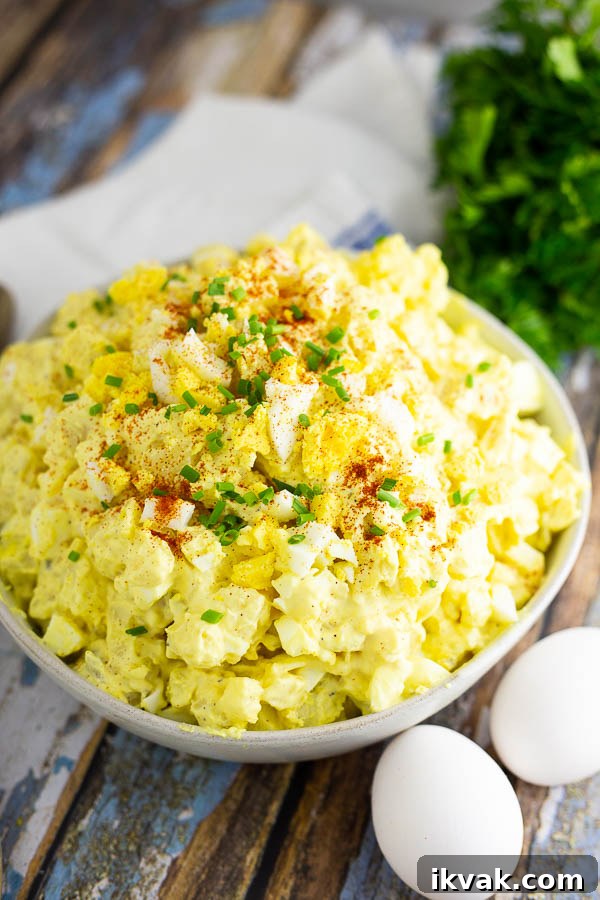 Deviled Egg Potato Salad Recipe — Creamy, Tangy Side Dish 2 Angle shot of deviled egg potato salad in a wide bowl topped with chives and paprika, next to 2 eggs and herbs on a rustic wooden background