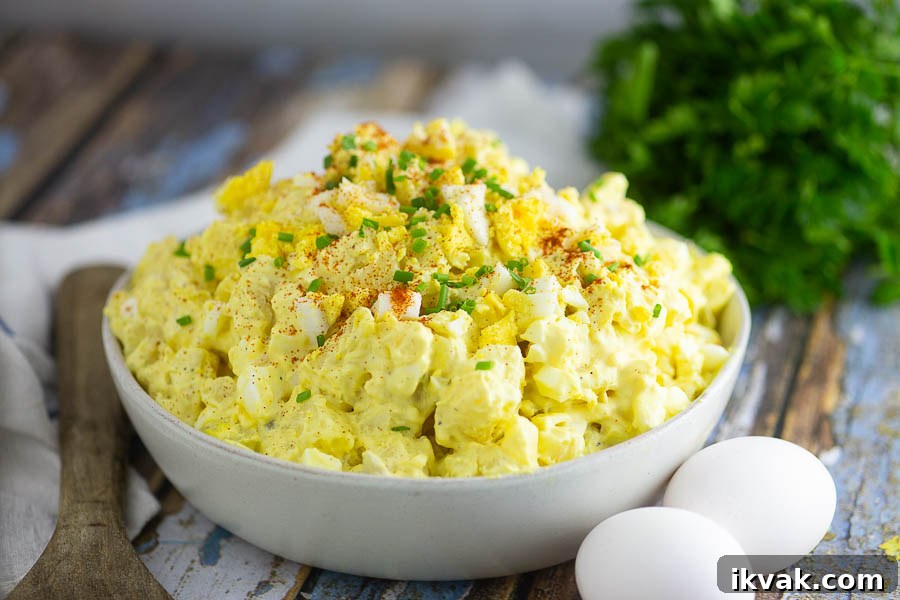 Deviled Egg Potato Salad Recipe — Creamy, Tangy Side Dish 3 Side shot of deviled egg potato salad in a bowl next to a wooden spoon, 2 eggs, and a bundle of herbs on a rustic wood background