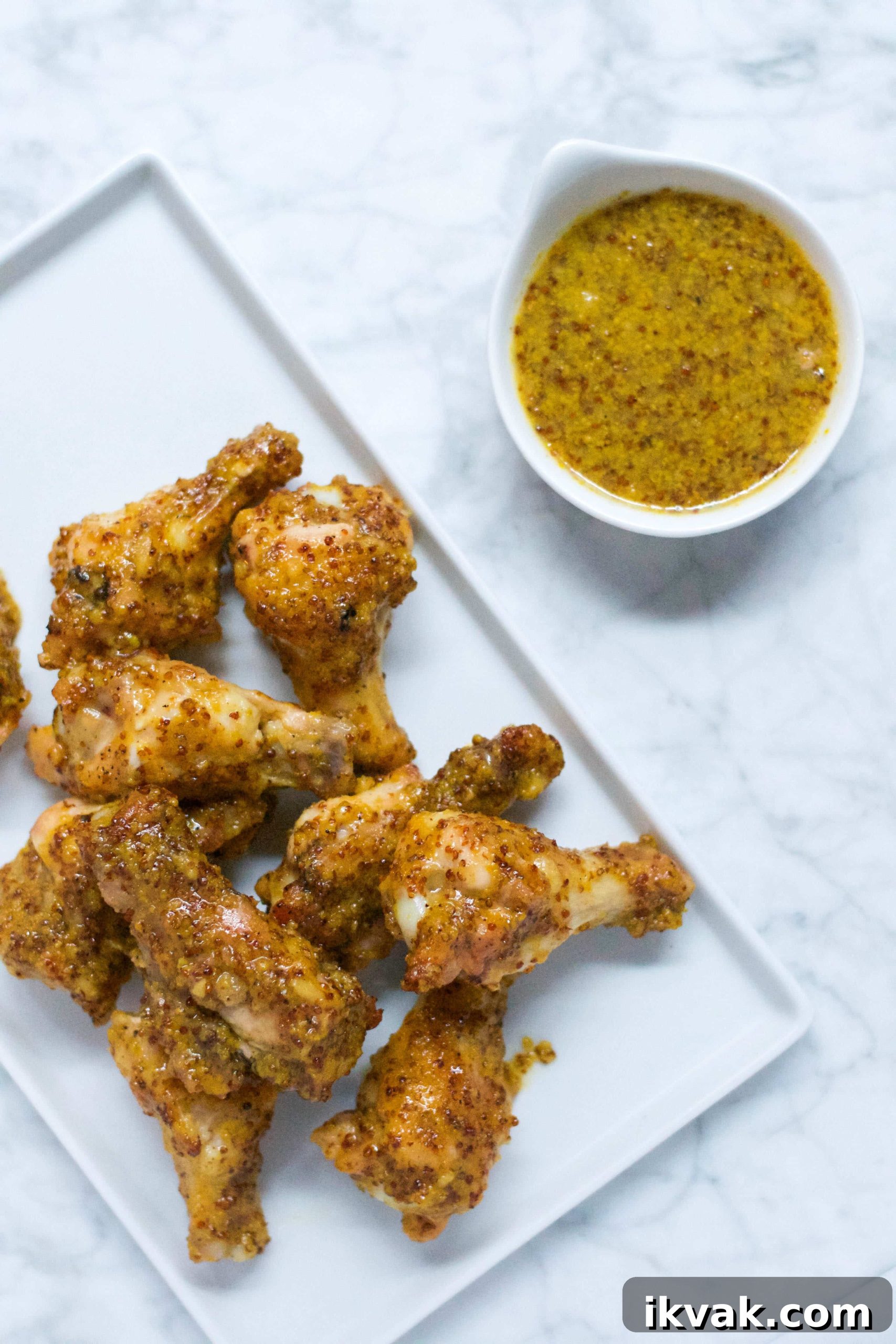 Crispy Honey Mustard Baked Chicken Wings Recipe 2 A rectangular white plate with several Honey Mustard Baked Chicken Wings, coated in a grainy mustard glaze, sits next to a small white bowl filled with mustard dipping sauce on a marble surface.