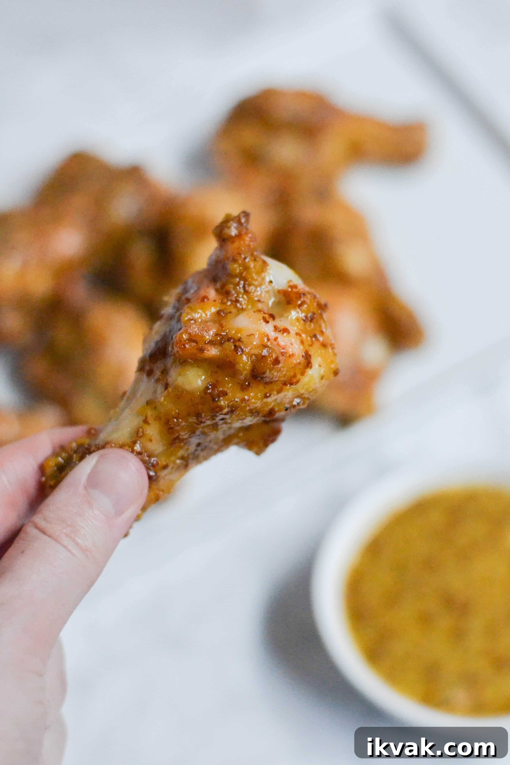 Crispy Honey Mustard Baked Chicken Wings Recipe 3 A hand holds a saucy Honey Mustard Baked Chicken Wing, with more wings blurred in the background and a small bowl of mustard dipping sauce nearby.