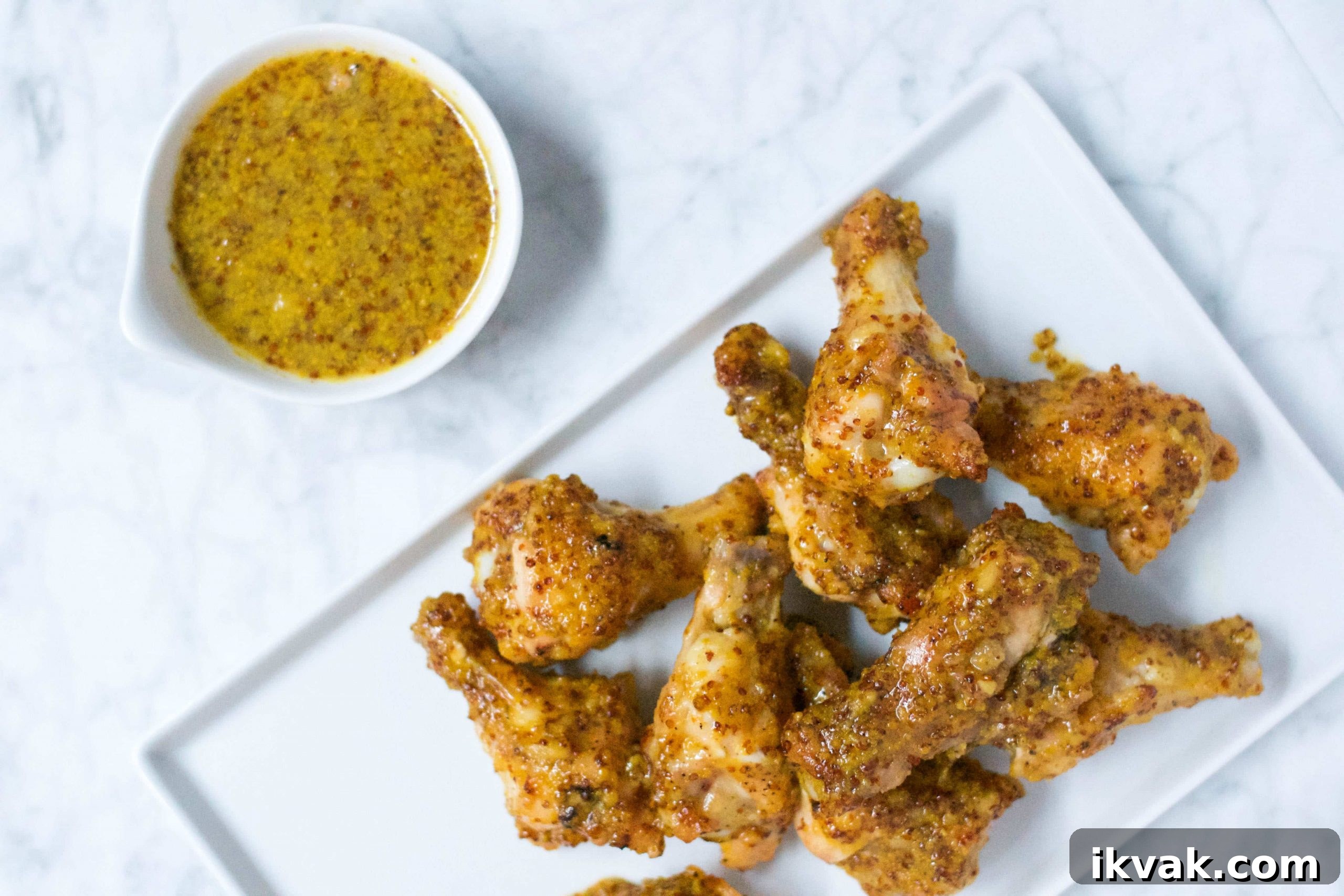 Crispy Honey Mustard Baked Chicken Wings Recipe 4 A white rectangular plate holds several pieces of crispy, Honey Mustard Baked Chicken Wings. Beside the plate, a small white bowl brims with grainy mustard dipping sauce, all set on a bright white marble surface.