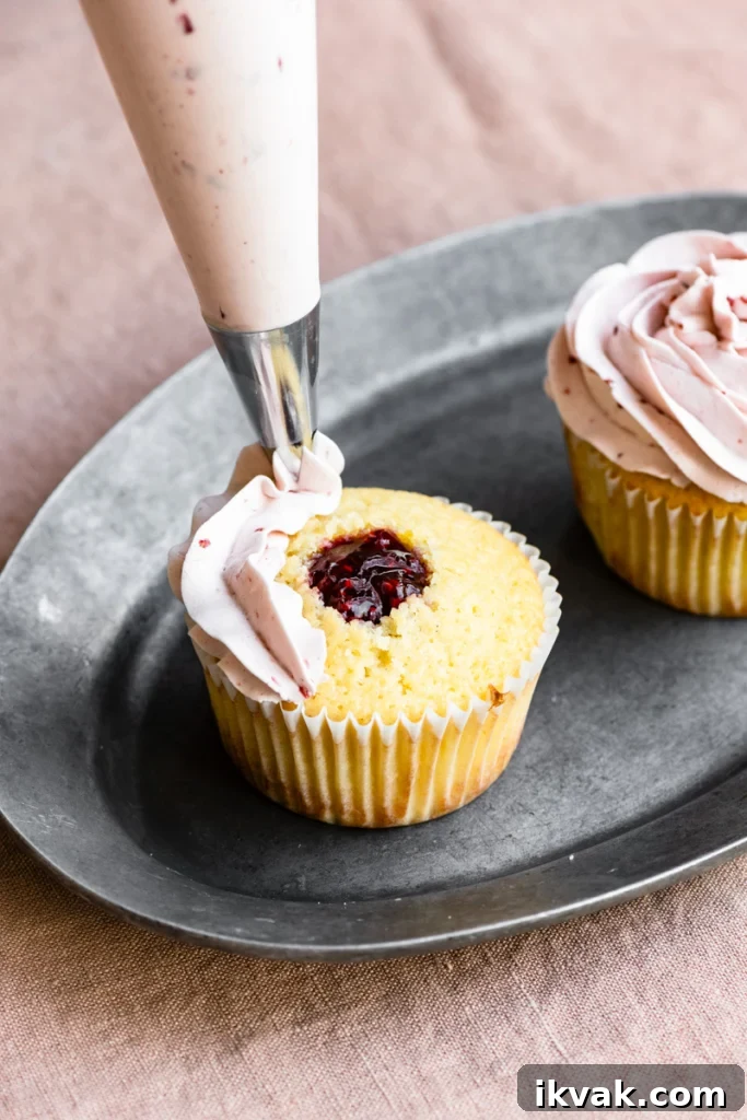 Piping bag starting to frost buttercream onto the top of a raspberry jam center filled cupcake.