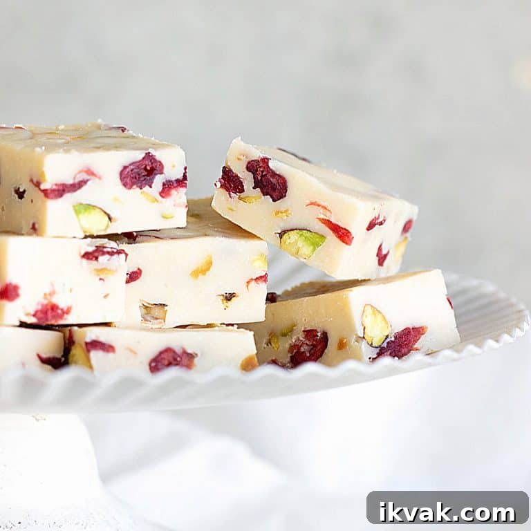 Several pieces of pistachio cranberry white chocolate fudge on a white cake stand with grey background.
