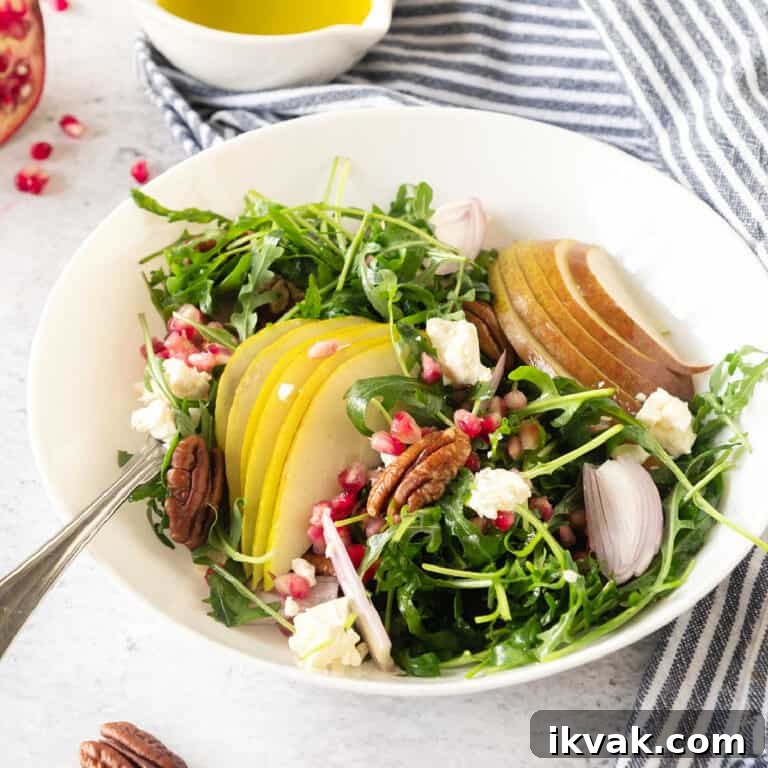 A white bowl with pear, arugula and pomegranate salad. Blue striped cloth. White surface.