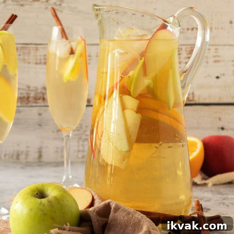 Tall pitcher and stem glasses of apple cider white wine sangria with sliced fruit. Whitish wood background.