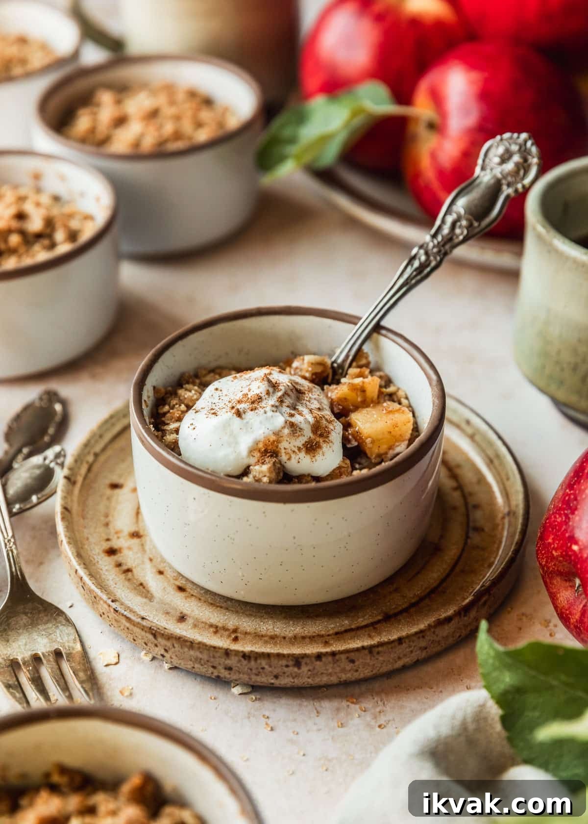 White ramekins with mini apple crumbles and oat topping on a tan counter next to apples, a tan linen, and two beige cups of coffee.