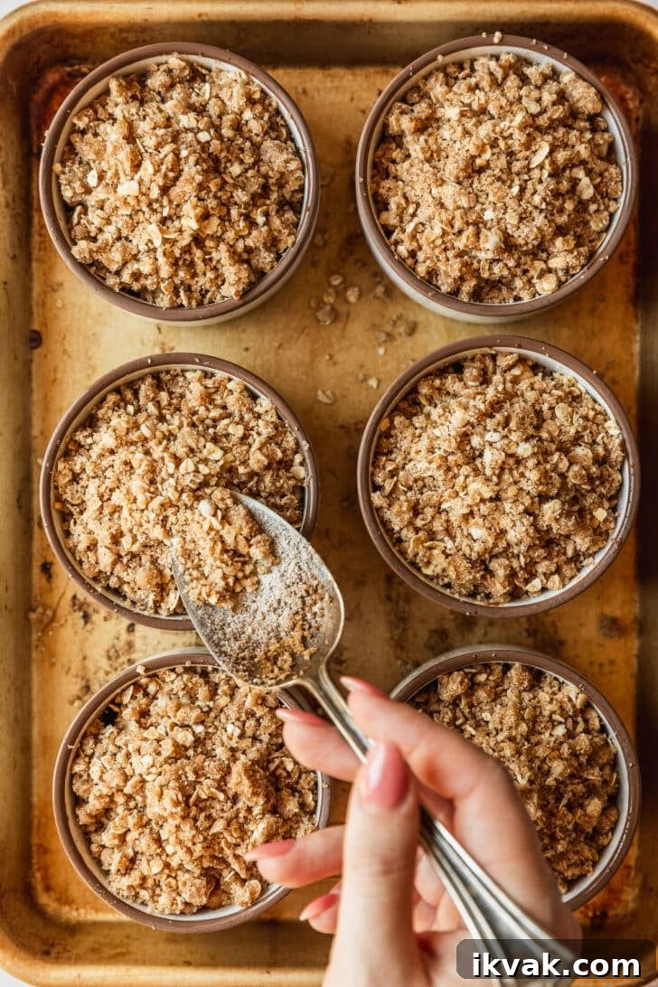 A hand spooning oat topping over mini apple crumbles on a gold pan.