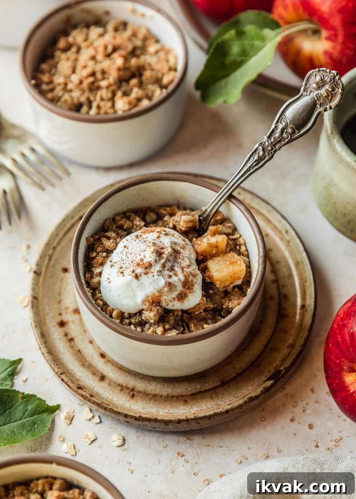 Mini apple crumbles in white ramekins on a tan counter next to beige cups of coffee, a beige linen, and apples.