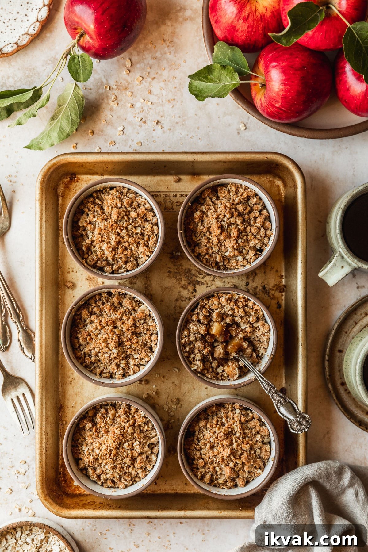 Six mini apple crumbles on a gold sheet pan next to beige cups of coffee, apples, a tan linen, and forks on a marble counter.