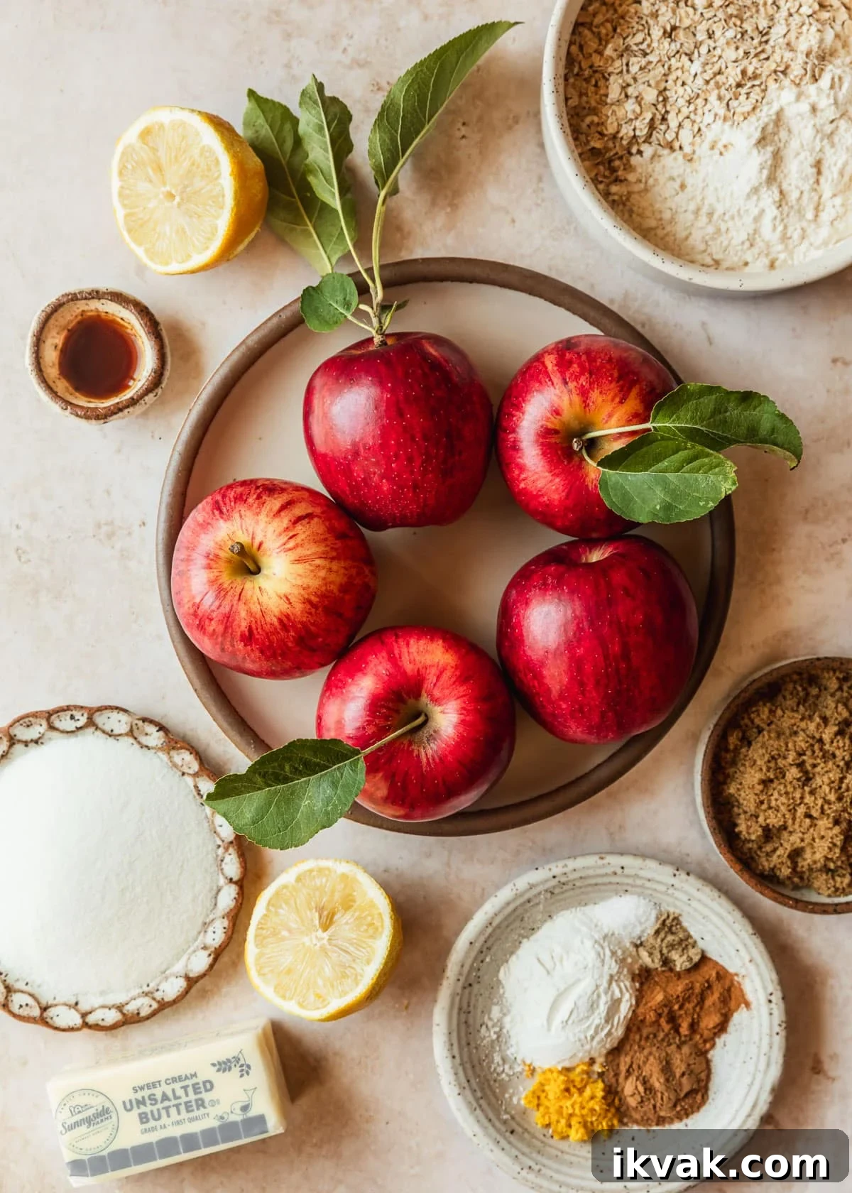 White bowls of honeycrisp apples, flour, oats, vanilla, sugar, brown sugar, and spices on a tan counter.