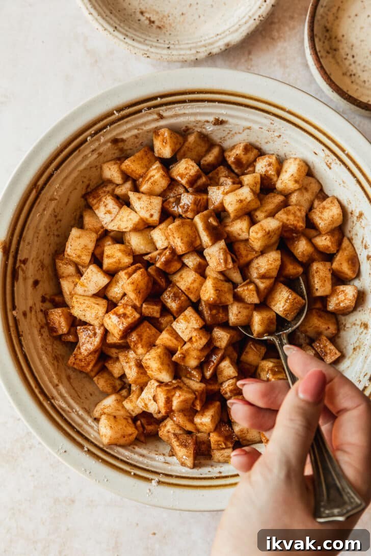A hand using a spoon to mix filling for mini apple crumbles in a white bowl.