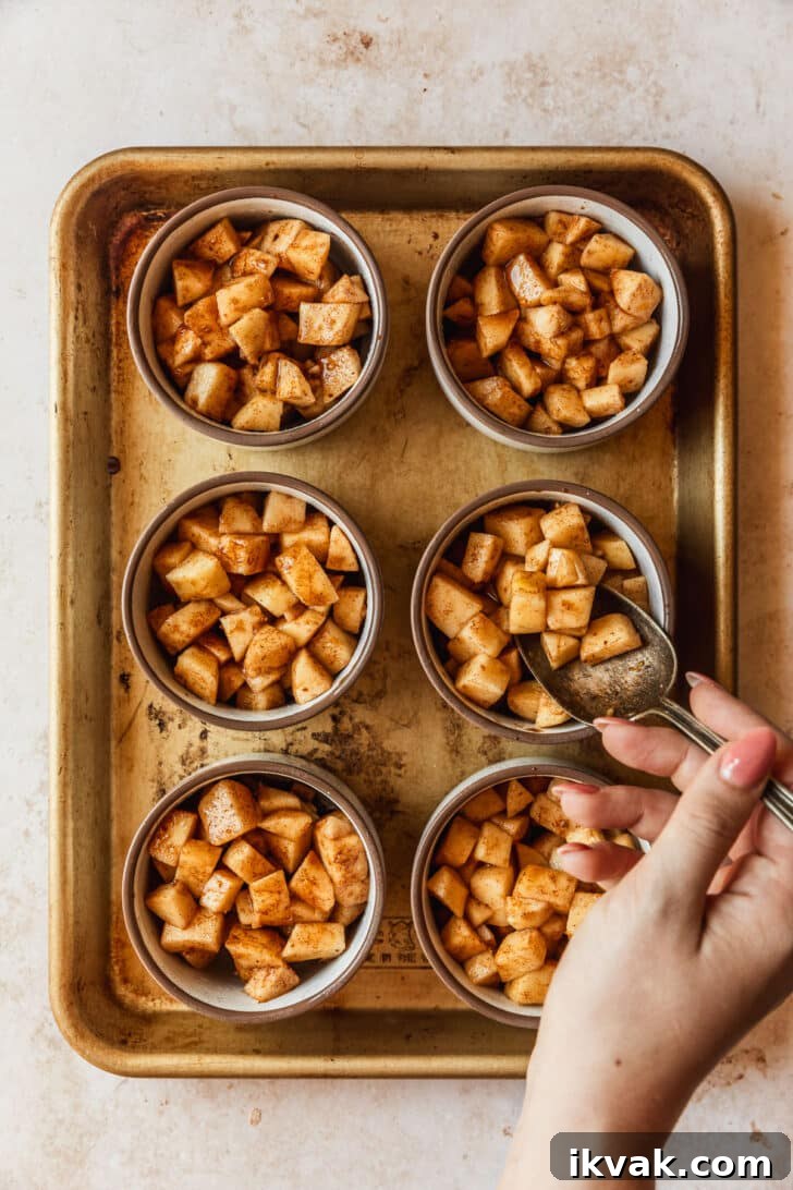 A hand spooning apple filling into ramekins on a gold sheet pan.